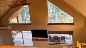 A room with a sloped ceiling, two large windows with a view of trees, a flat-screen TV mounted on the wall, and a wooden ceiling beam. - Secluded 3 bed house in Kila with flowing creek, hot tub, with plenty of parking (Kila)