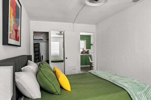 Ett sovrum med en säng, kuddar, en grå soffa, ett grönt överkast, en vit dörr som leder till ett kök och en inramad tavla på väggen.