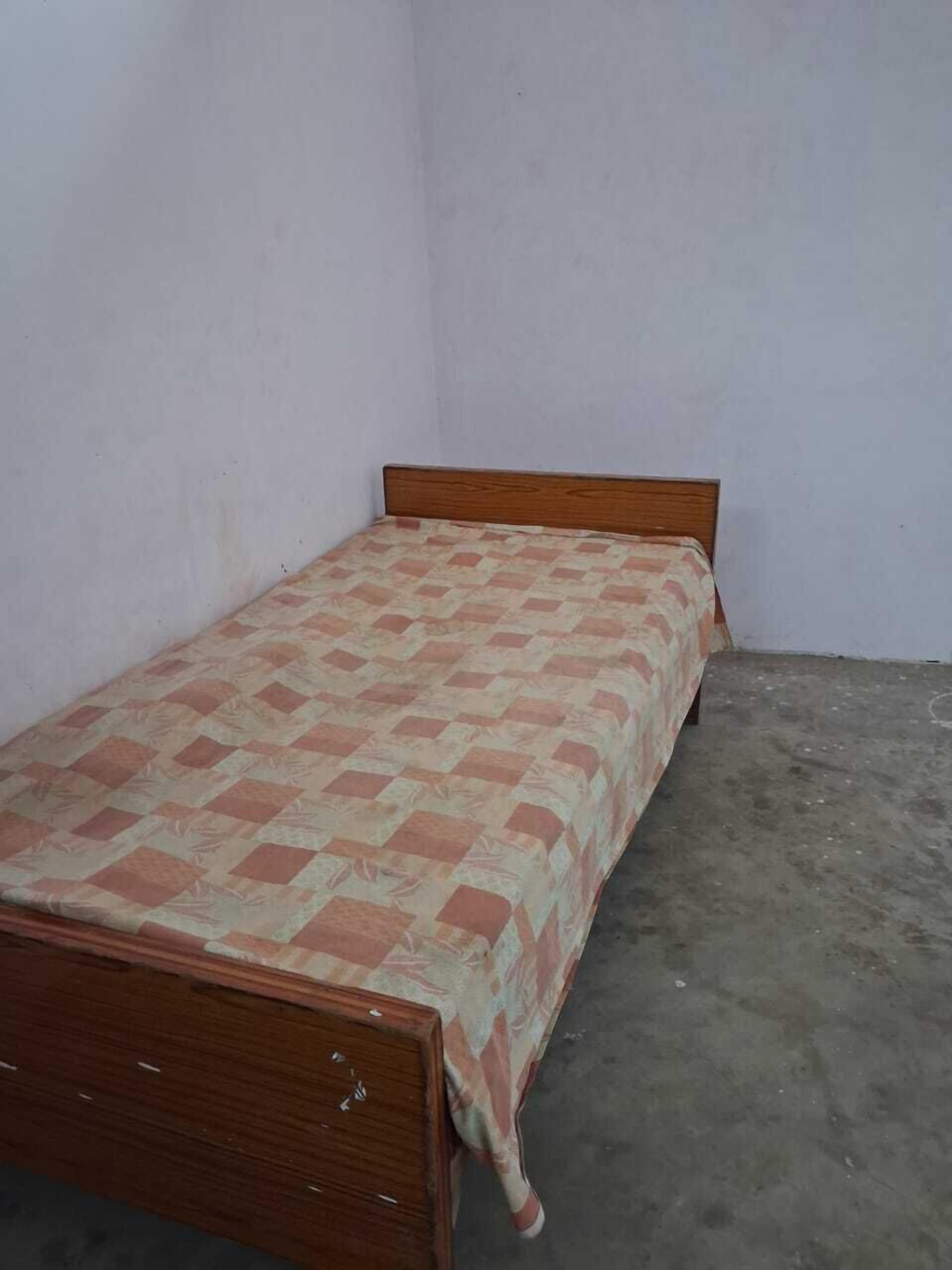 Una cama individual con estructura de madera y colcha estampada en una habitación con paredes lisas y suelo de concreto.