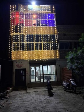A multi-story building with illuminated string lights, a ground-level entrance, and parked scooters. - Athithi Homestay (Jaunpur)