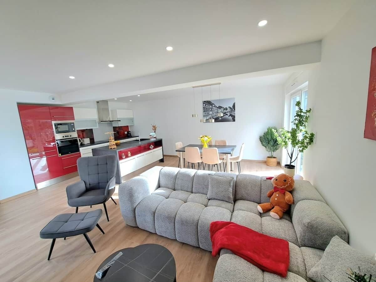 A modern living room with a grey sectional sofa, a grey armchair, and a small ottoman. There is a dining area with a table and chairs, and a kitchen in the background.
