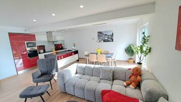 A modern living room with a grey sectional sofa, a grey armchair, and a small ottoman. There is a dining area with a table and chairs, and a kitchen in the background.