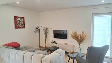Een moderne woonkamer met een hoekbank, een grijze fauteuil, een kleine ronde tafel, een flatscreen televisie en een vaas met gedroogde planten.