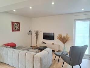 A modern living room with a sectional sofa, a grey armchair, a small round table, a flat-screen TV, and a vase with dried plants. - Chez Fernand - New home in Colmar with parking and a garden (Colmar)