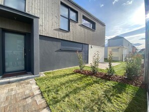 Een modern, twee verdiepingen hoog gebouw met een donkere gevel, een glazen entree en een verzorgd gazon met kleine struiken.