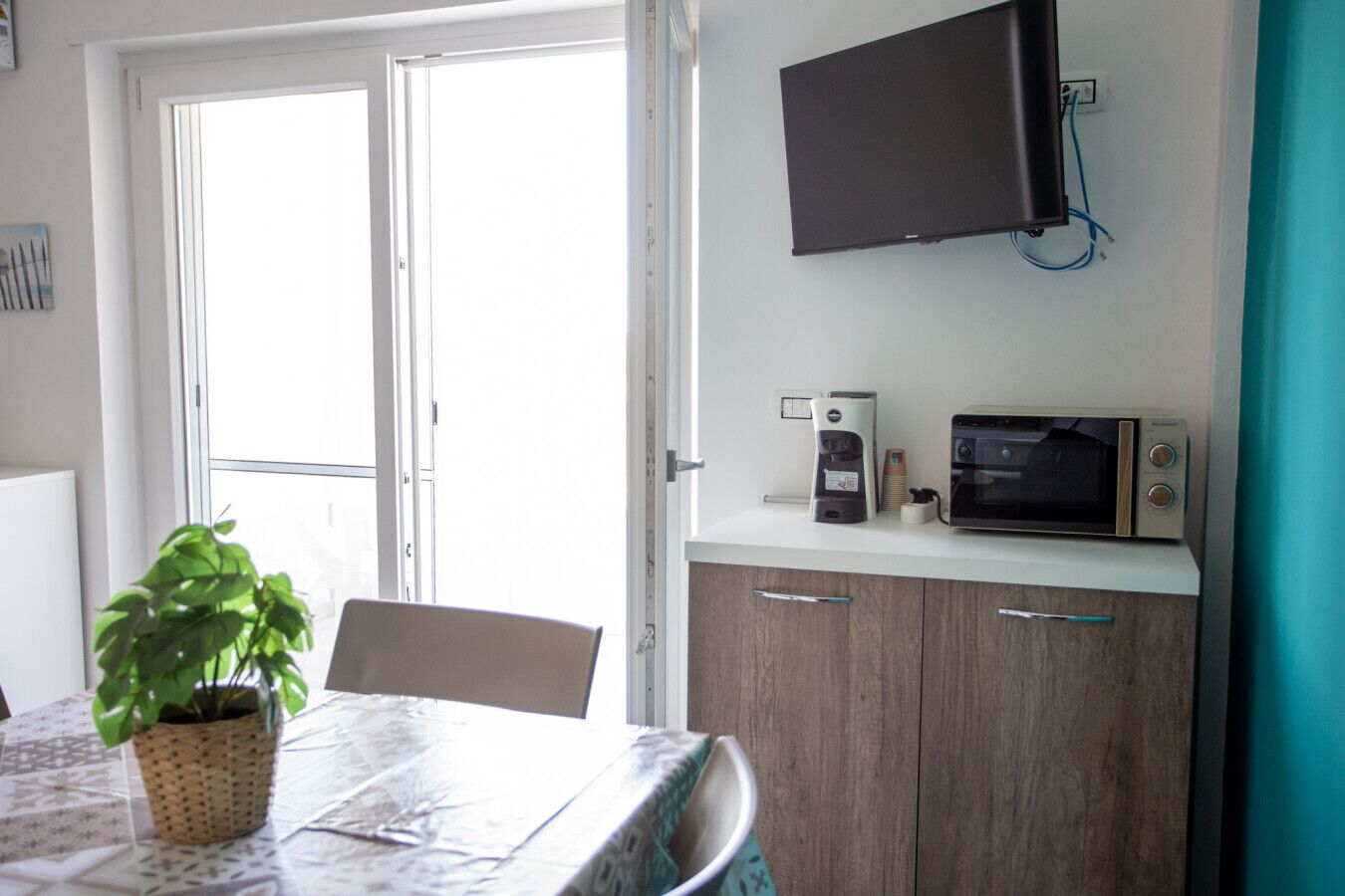 A room with a dining table, chairs, a potted plant, a microwave, a coffee maker, and a TV mounted on the wall.