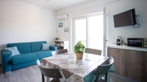Una sala de estar moderna con un sofá azul, una mesa de comedor con mantel estampado y una zona de cocina con microondas y cafetera.