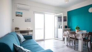 Una sala de estar moderna con un sofá color turquesa, una mesa de comedor con sillas, y una cocina visible a través de una puerta abierta.