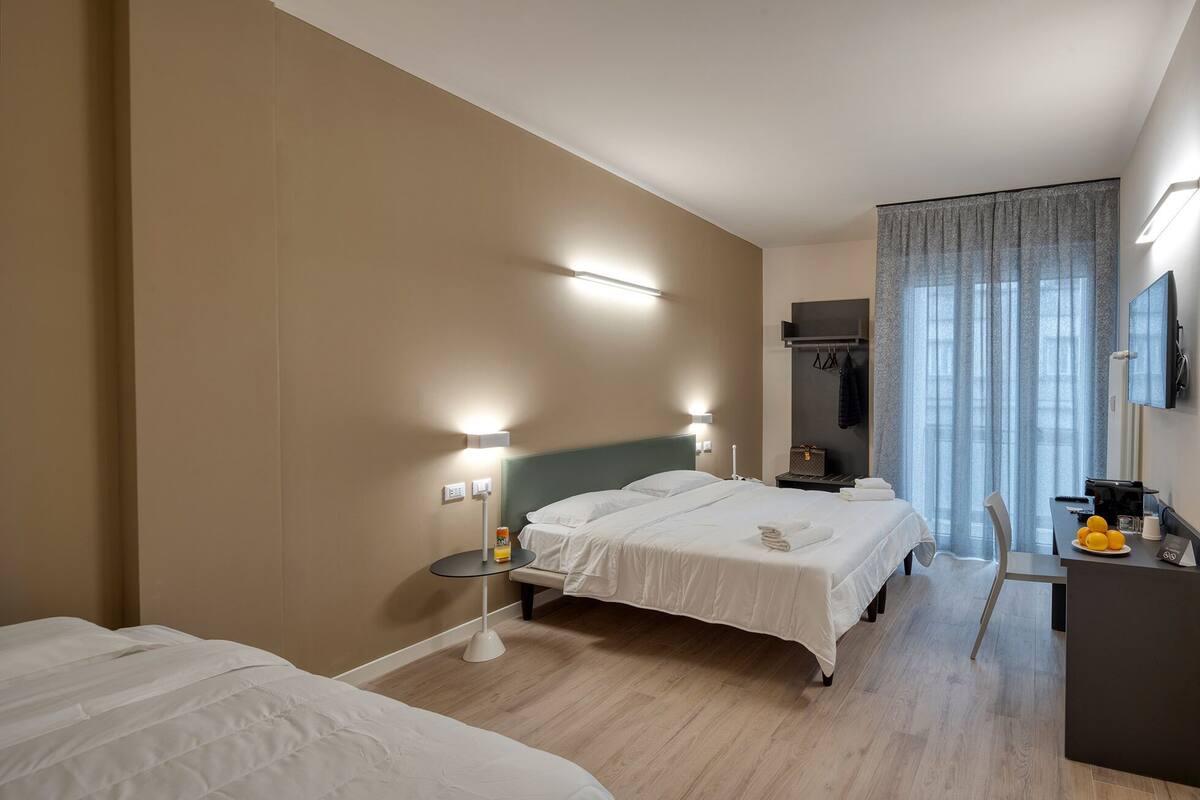Une chambre d’hôtel avec deux lits, un bureau avec une chaise, un minibar et une grande fenêtre avec des rideaux.
