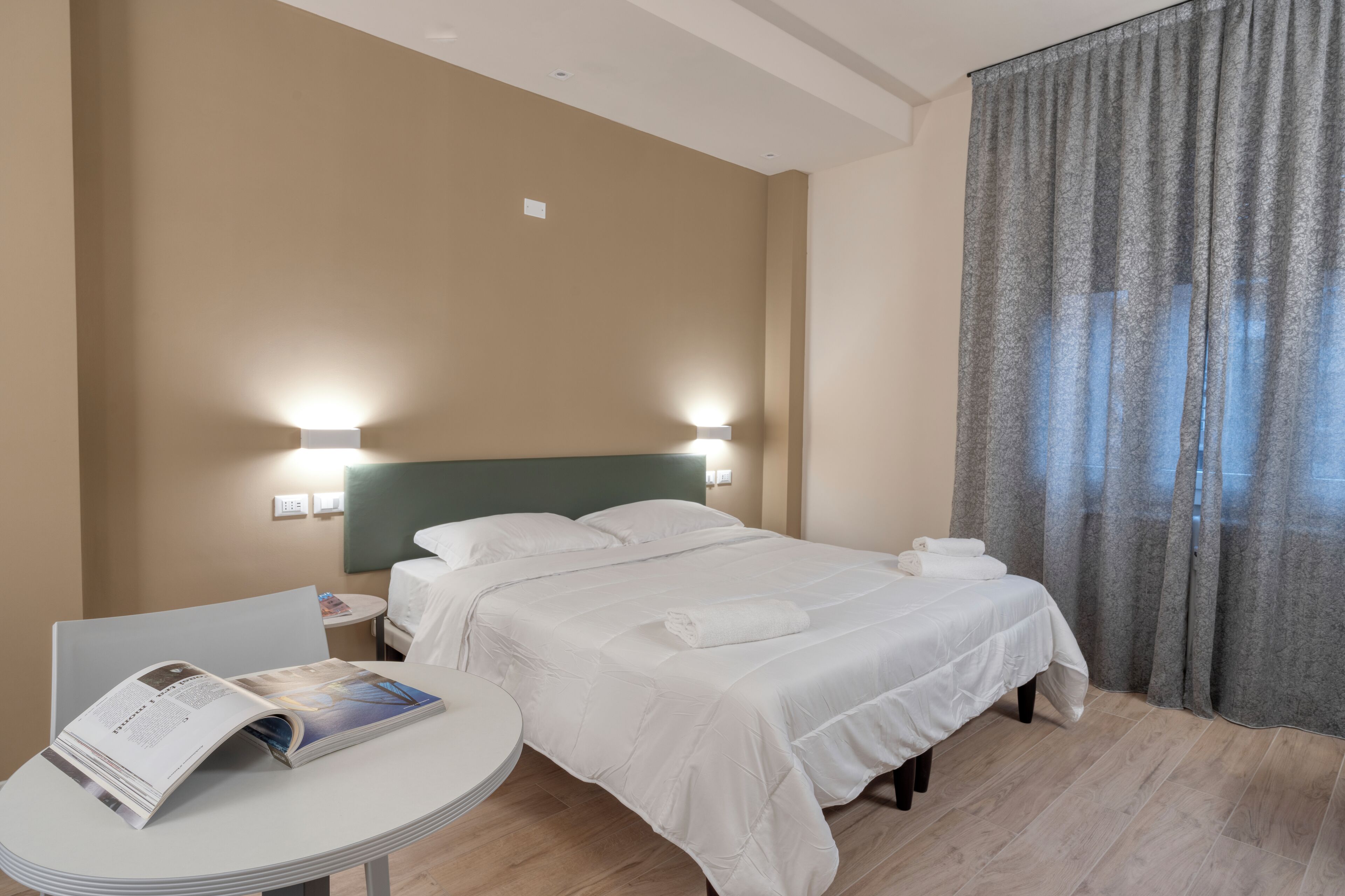 Een hotelkamer met een bed, een nachtkastje met een tijdschrift en een raam met gordijnen.