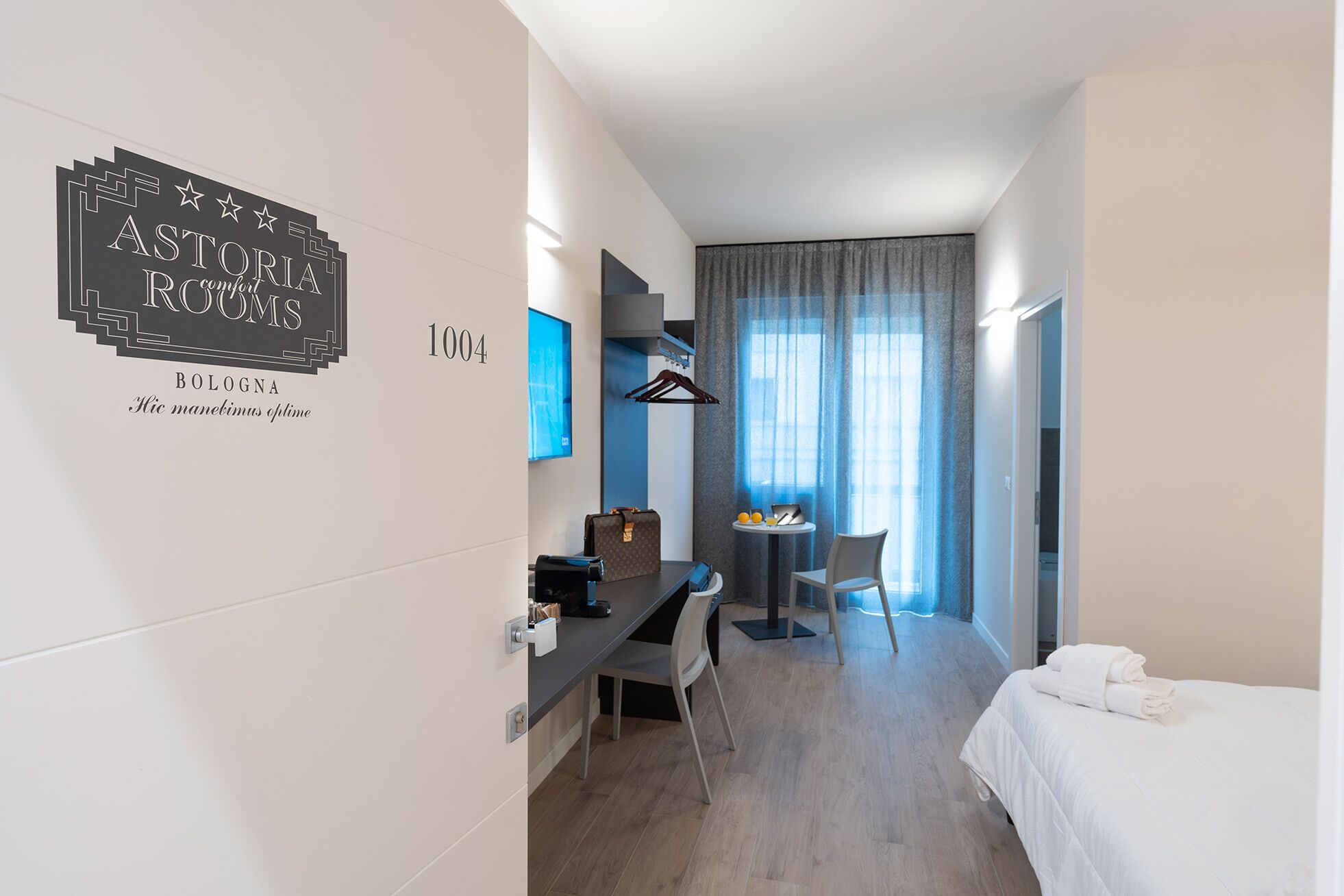 A hotel room with a bed, a desk, a chair, a small table with a laptop and oranges, and a window with curtains.