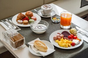 Una tavola imbandita per la colazione con croissant, fragole, macarons, una tazza di caffè, un bicchiere di succo d'arancia, uova strapazzate, salsicce e una piccola ciotola di yogurt.