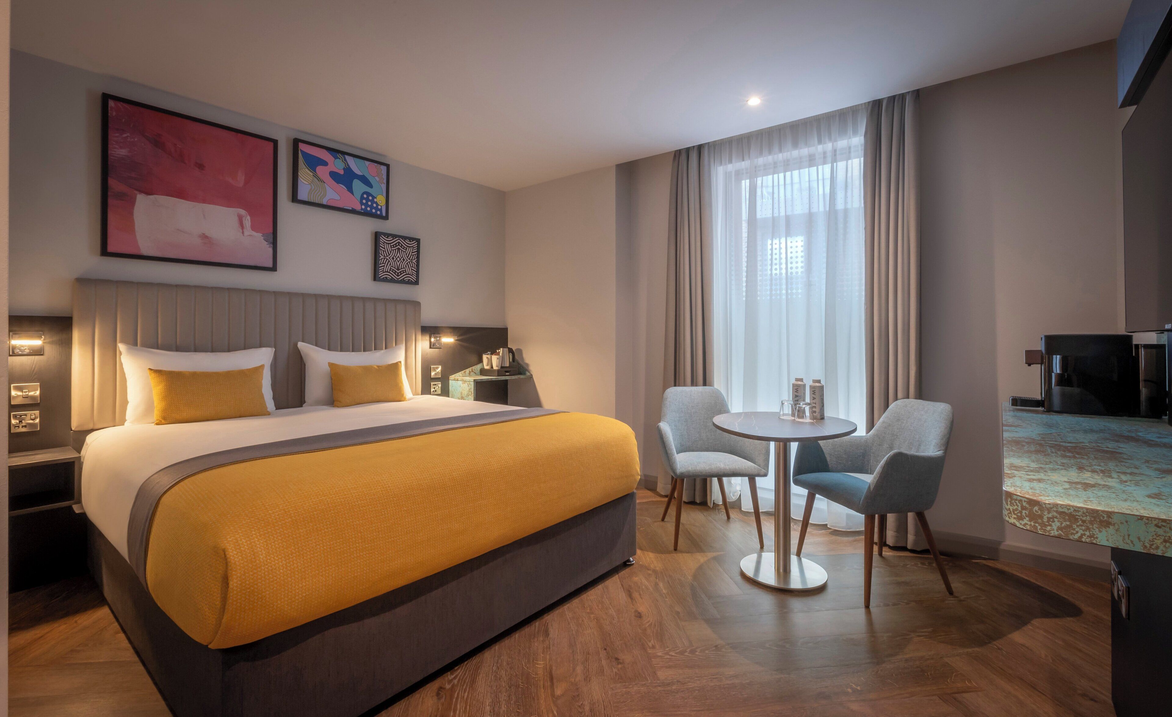 Habitación de hotel con una cama grande, una mesa redonda con sillas, televisión y obras de arte en las paredes.