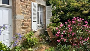 Una casa de piedra con una puerta y contraventanas blancas, un banco de madera y un jardín con diversas plantas y flores.