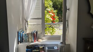 Una ventana con cortinas translúcidas, una estantería con libros y objetos decorativos, un tostador, una cesta con pan, y una mesa con una bandeja de platos.