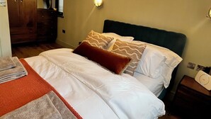 Ein ordentlich bezogenes Bett mit weißen Leinen und gemusterten Kissen, ein Holzschrank, eine wandmontierte Lampe und ein kleiner Nachttisch mit einer Uhr.