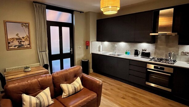 Ein modernes Wohnzimmer mit einer braunen Ledersofa, einem kleinen Holztisch und einer Fernbedienung darauf. Der Raum hat Holzböden und ein großes Fenster mit Vorhängen.