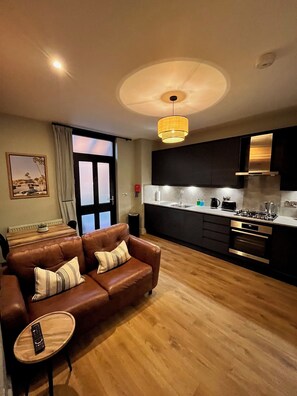A modern living room with a brown leather sofa, a small wooden side table, and a remote control on it. The room has wooden flooring and a large window with curtains. - Beach Retreat | Stylish 1-Bed in Canford Cliffs (Poole)