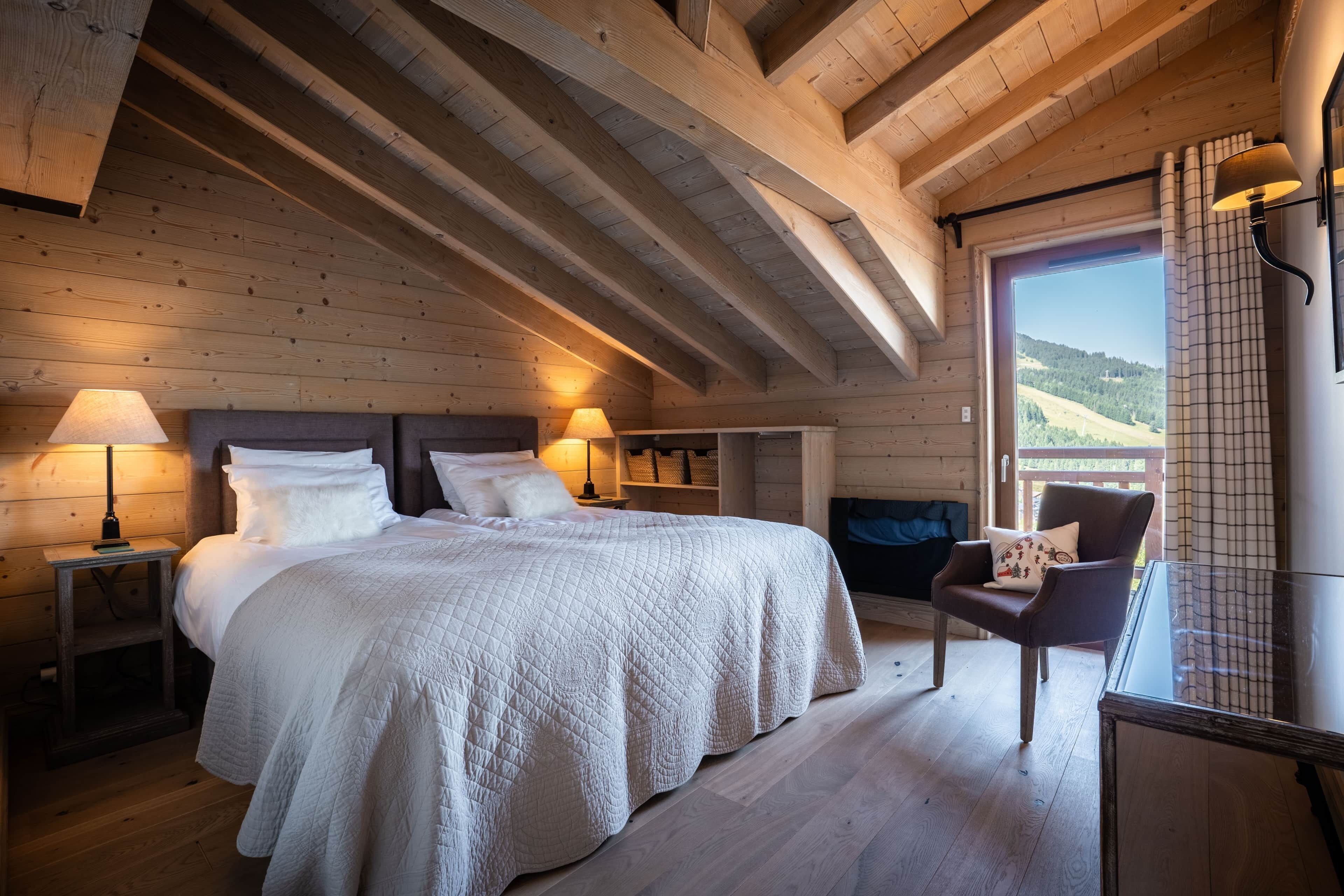 A bedroom with a large bed, wooden ceiling beams, a chair, and a view of the outdoors.