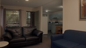 Un soggiorno con un divano in pelle nera, un divano blu con motivi decorativi e un tavolino rotondo. Sullo sfondo, si intravede una cucina con un forno a microonde e degli armadietti.