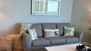 Un salon moderne avec un canapé gris, des coussins à motifs, une œuvre d’art encadrée et une table basse blanche.