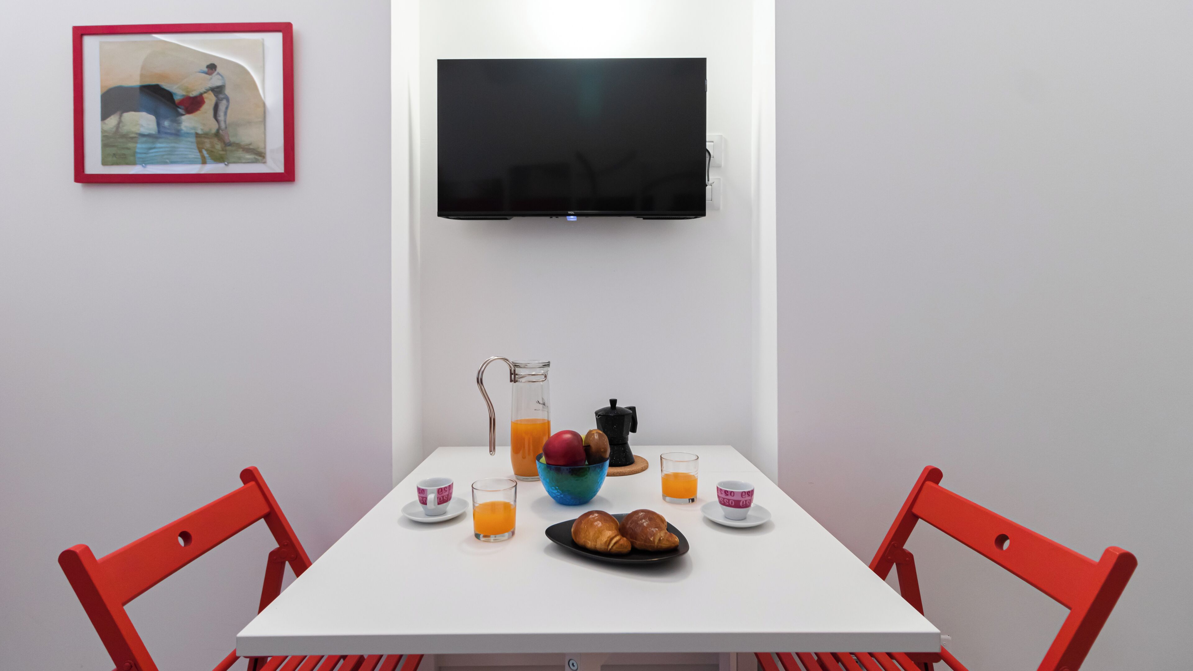 A white table with a television above it, a pitcher of orange juice, a coffee maker, a bowl of fruit, and two croissants on a white tablecloth.