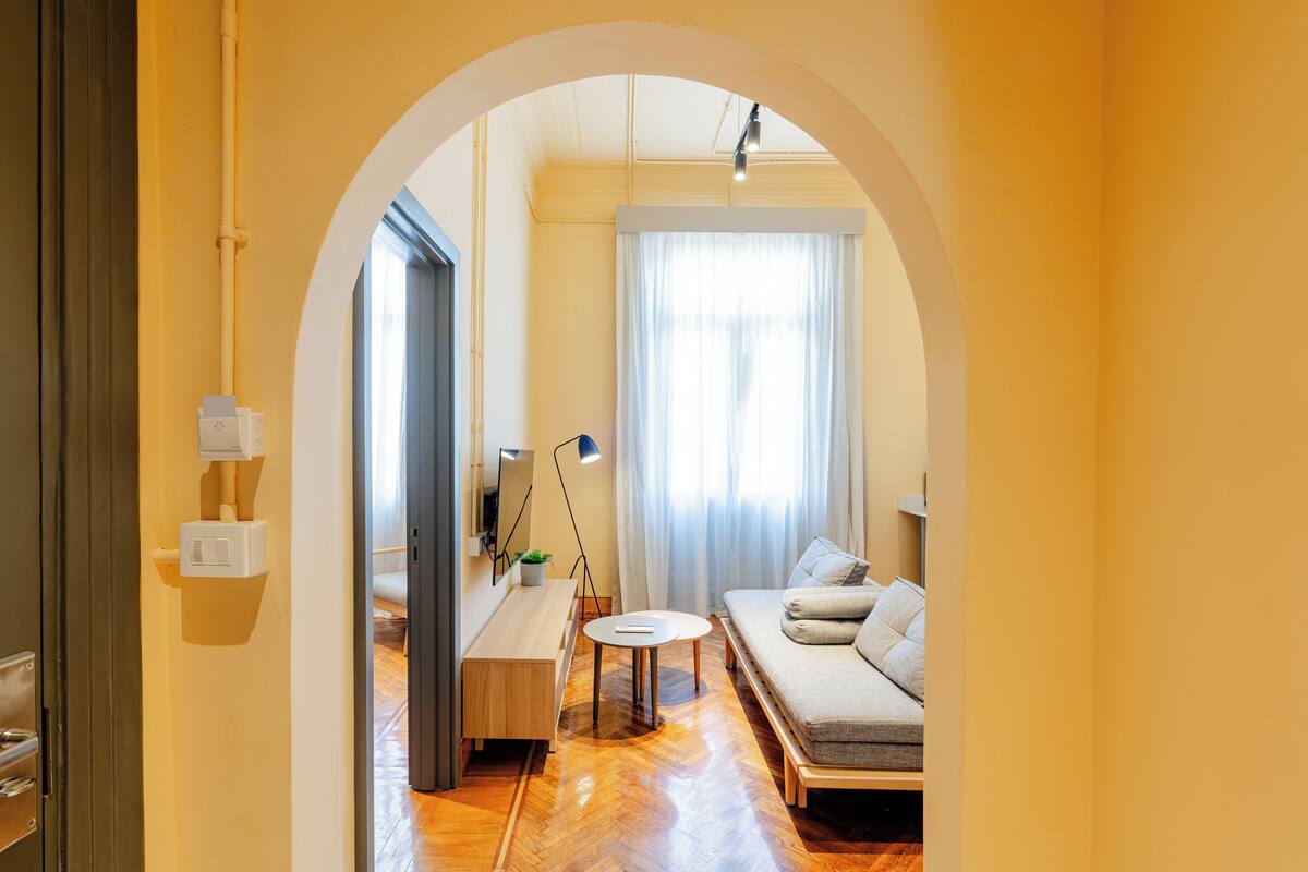 Una sala de estar moderna con un sofá, una mesa de centro y una lámpara. El suelo es de madera y hay un ventanal con cortinas.