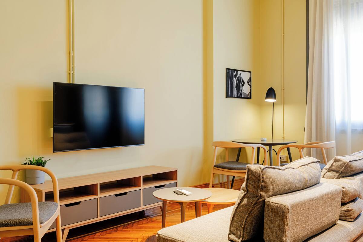 Una sala de estar moderna con un televisor de pantalla plana, muebles de madera, una pequeña mesa redonda y un sofá con cojines.