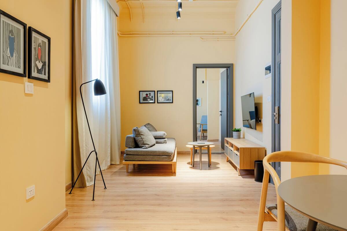 Un moderno salón con un sofá, una pequeña mesa redonda y una lámpara de pie. Hay un espejo en la pared y un televisor montado sobre un mueble de madera.