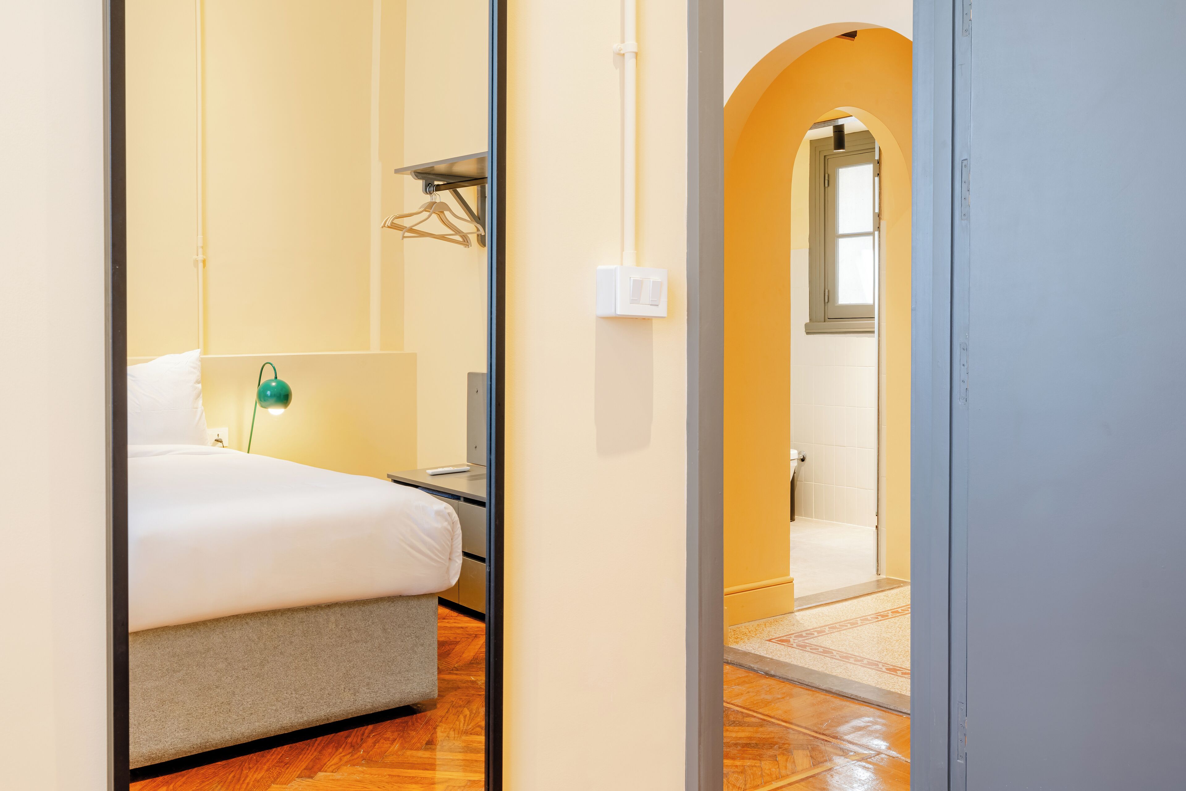 A hotel room with a bed, bedside table, lamp, and a view of the hallway and bathroom.