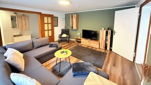 A modern living room with a grey sofa, wooden flooring, a small yellow table, and a flat-screen TV. - Cozy vacation apartment in Nideggen-Berg (Nideggen)
