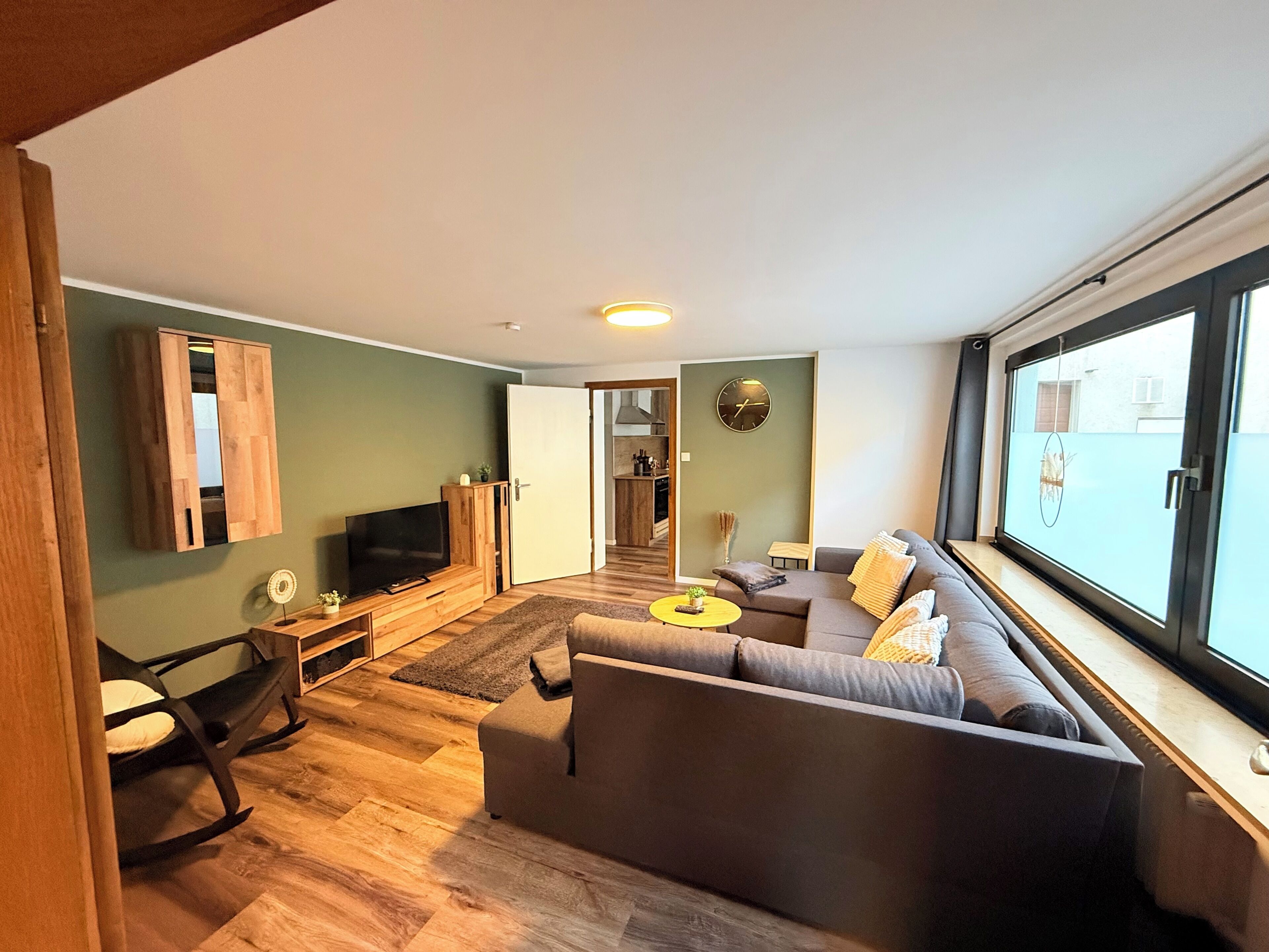 Un salon moderne avec un canapé, un téléviseur et une vue dégagée sur l’extérieur grâce à une grande fenêtre.