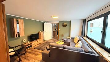 Un salon moderne avec un canapé, un téléviseur et une vue dégagée sur l’extérieur grâce à une grande fenêtre.