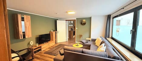 Un salon moderne avec un canapé, un téléviseur et une vue dégagée sur l’extérieur grâce à une grande fenêtre.