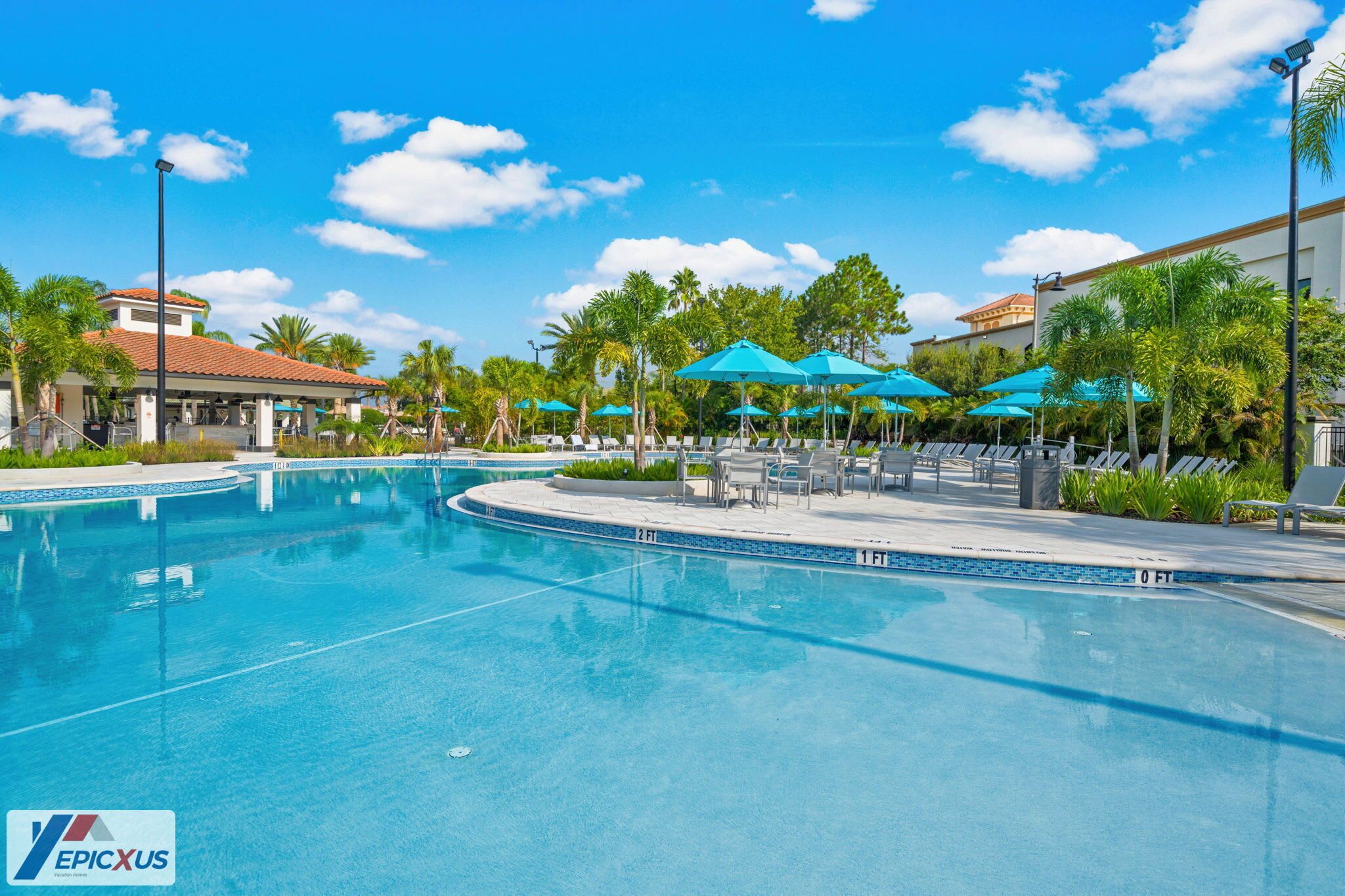A swimming pool with lounge chairs and umbrellas, surrounded by palm trees and a building with a red roof.