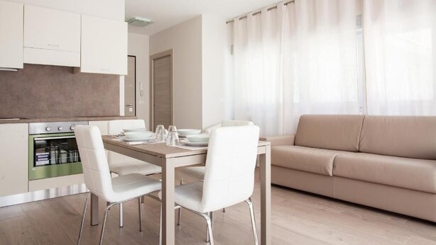 Una cocina moderna con armarios blancos, una mesa de comedor con sillas y un sofá beige en una sala de estar con un ventanal y cortinas translúcidas.