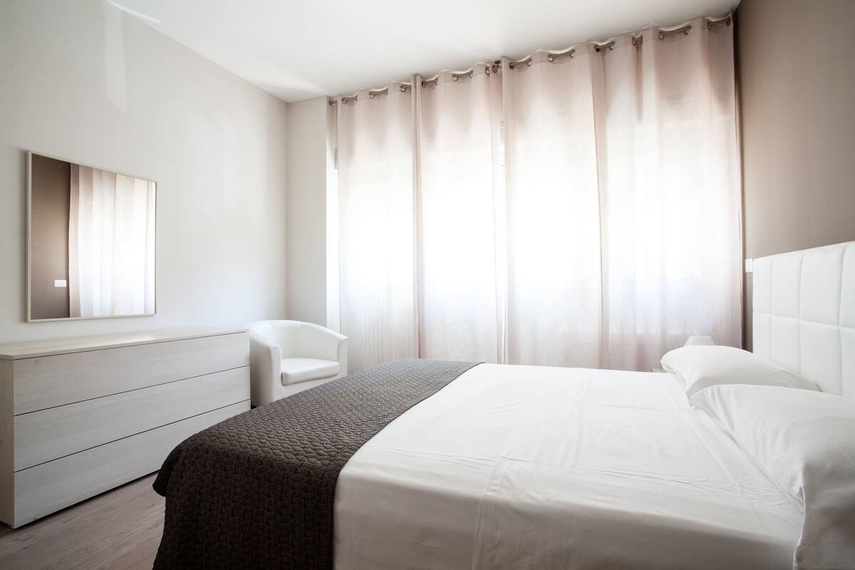 Habitació d'hotel moderna amb un llit gran, una taula de nit, una cadira i un armari.