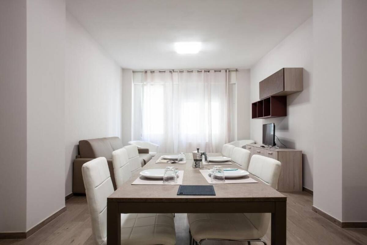 Zona de menjador moderna amb una taula de fusta preparada per a quatre, envoltada d'escons blancs. Un televisor està penjat a la paret a la dreta.