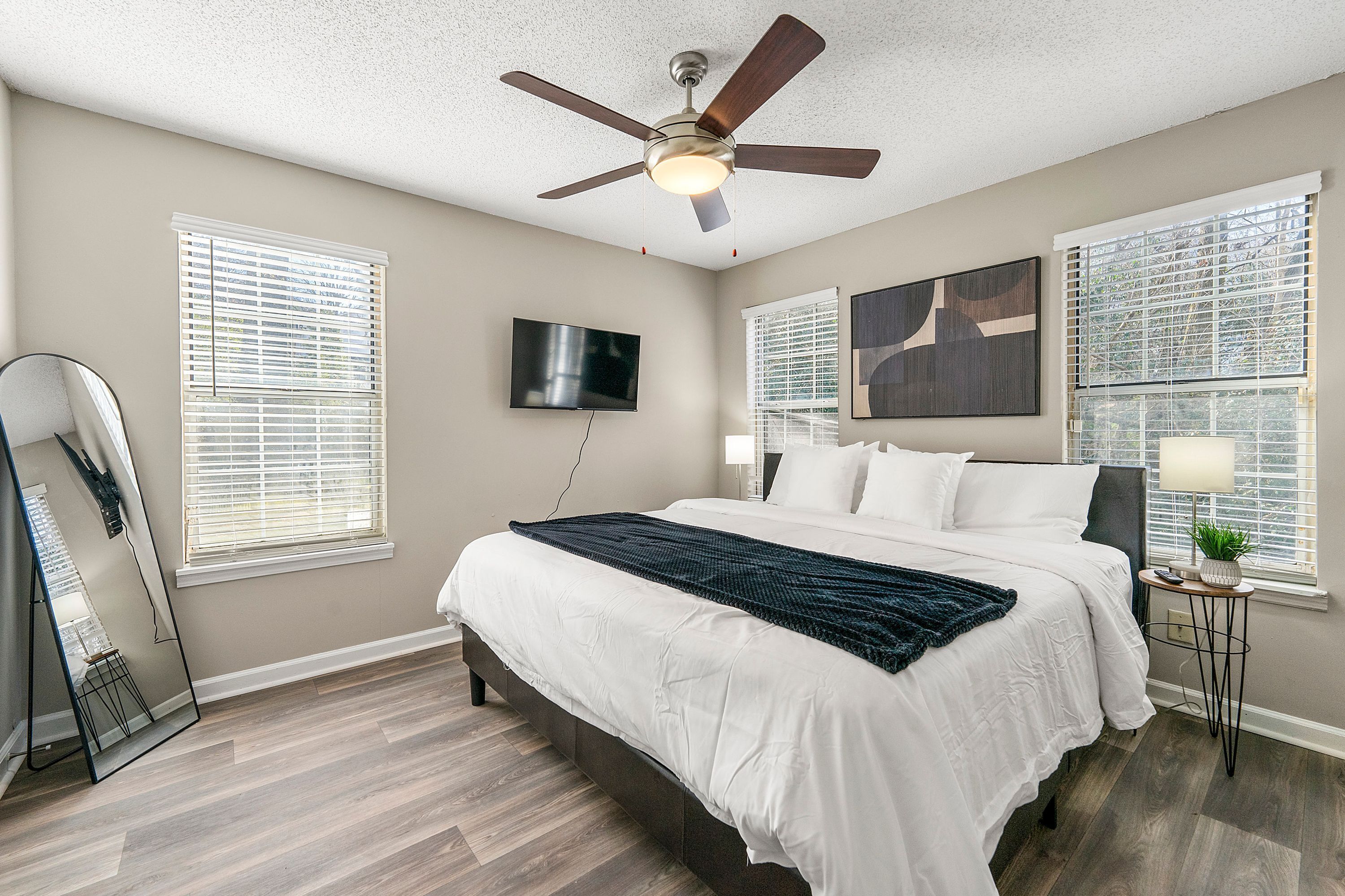 A bedroom with a bed, ceiling fan, TV, and windows with blinds.