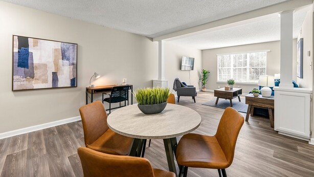 Een ruime woonkamer met een ronde tafel, twee oranje fauteuils, een grijze bank, een bijzettafel met een plant en een groot abstract schilderij aan de muur.