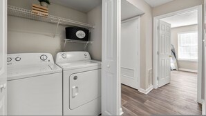 Een wasserij met een voorlader wasmachine en droger, een plank met een potplant, en een deur naar een andere kamer.