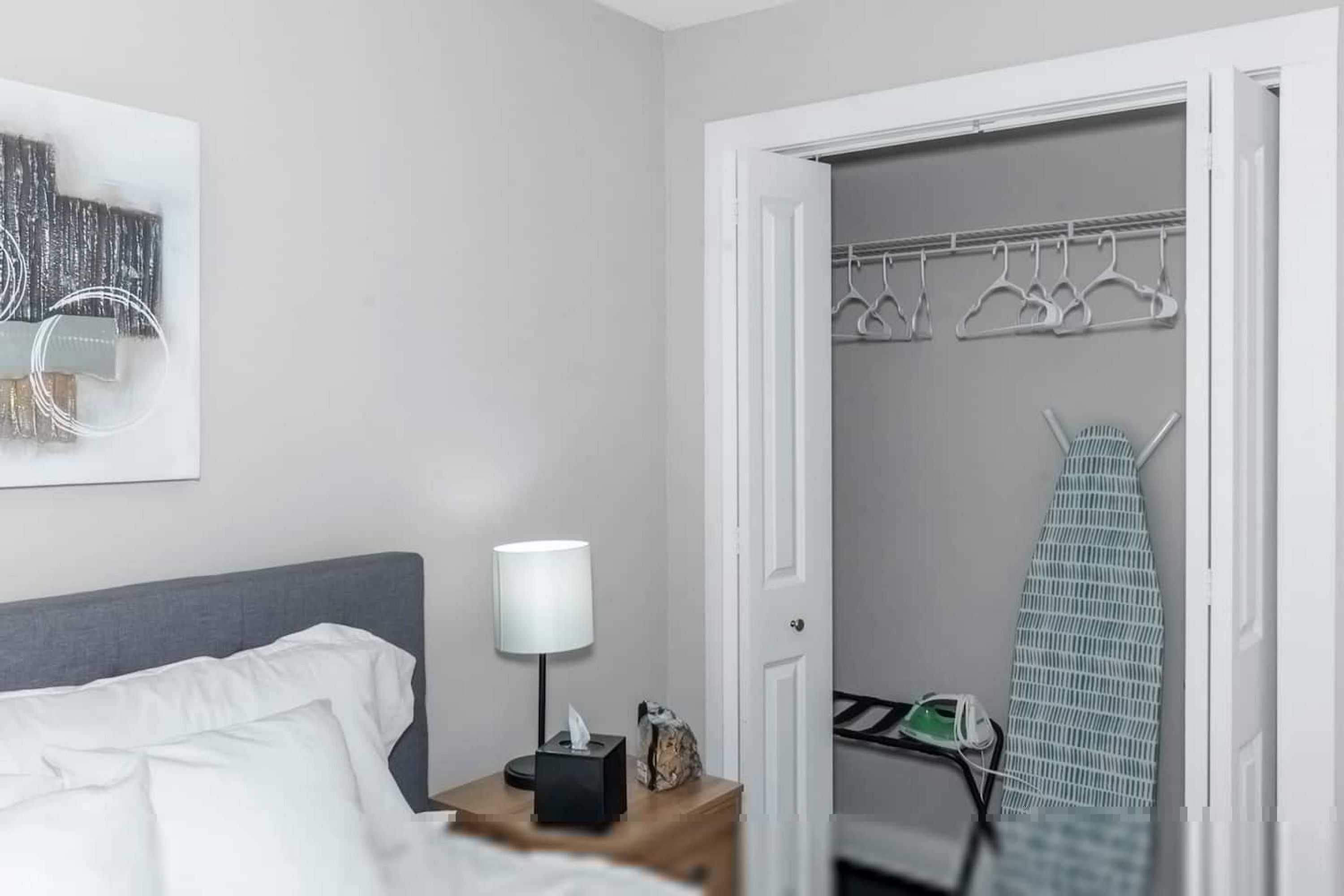 Habitación de hotel con una cama bien hecha, una lámpara de noche, una mesita con una caja de pañuelos y un adorno, un perchero con ganchos y una plancha con tabla de planchar.