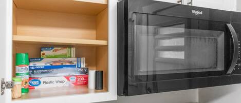 Een keuken met een zwarte magnetron en een witte kast eronder met verschillende keukenbenodigdheden.