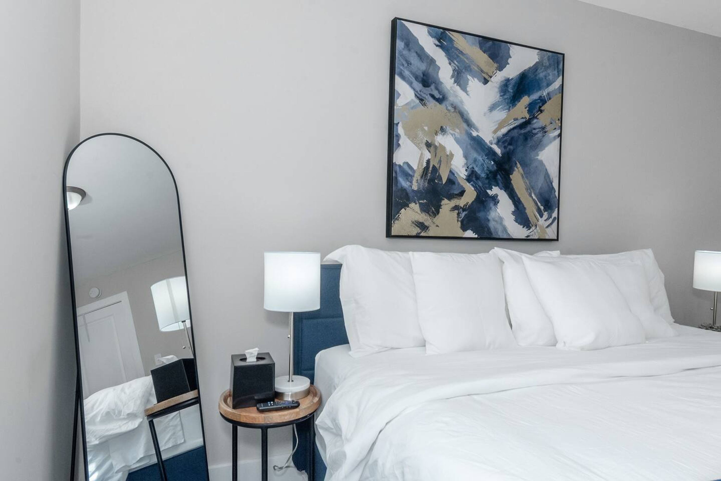 A hotel room with a large bed, two bedside lamps, a bedside table with a lamp, a mirror, and a painting on the wall.