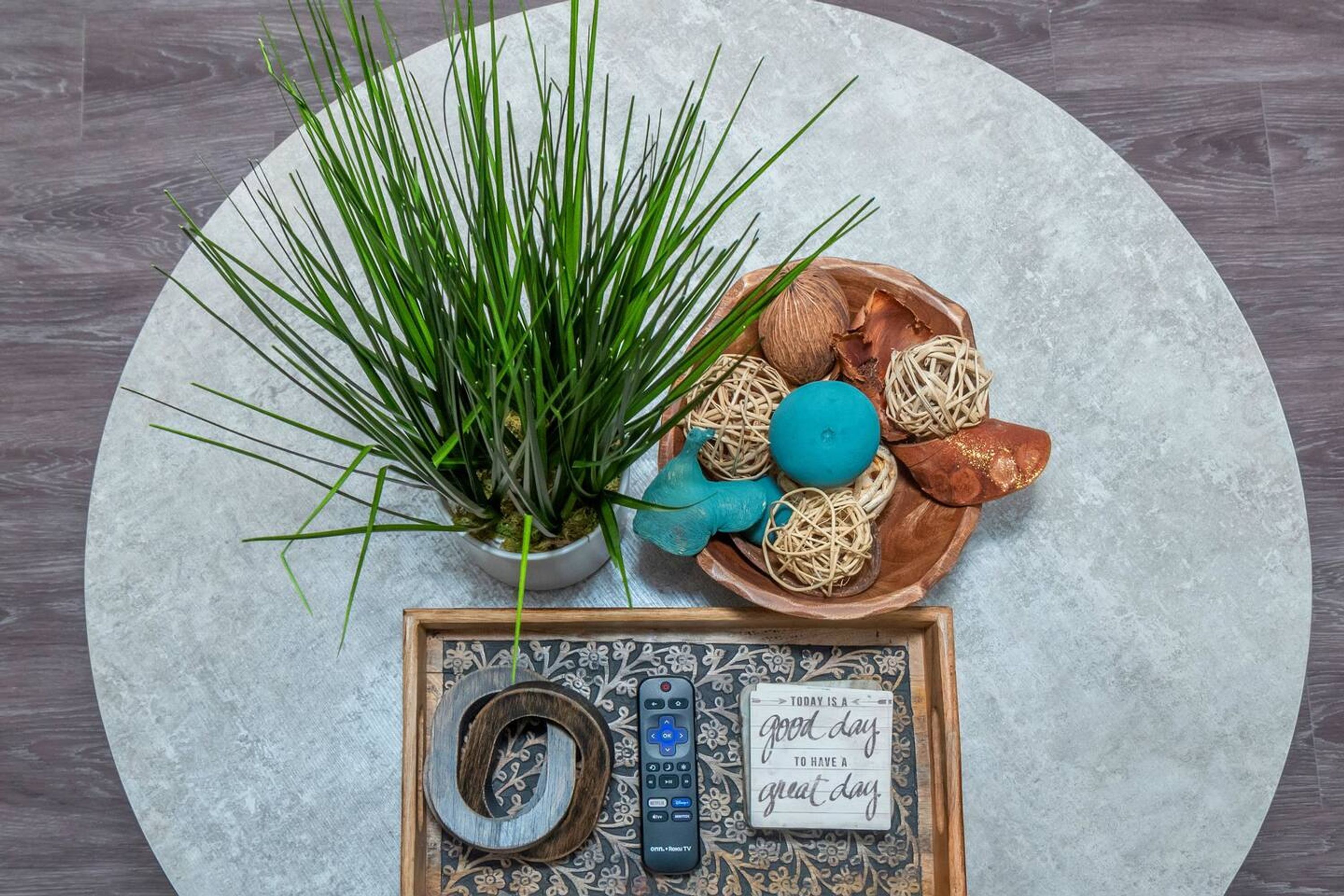 A round table with a potted plant, a bowl of decorative items, and a tray with a remote control and a framed card.