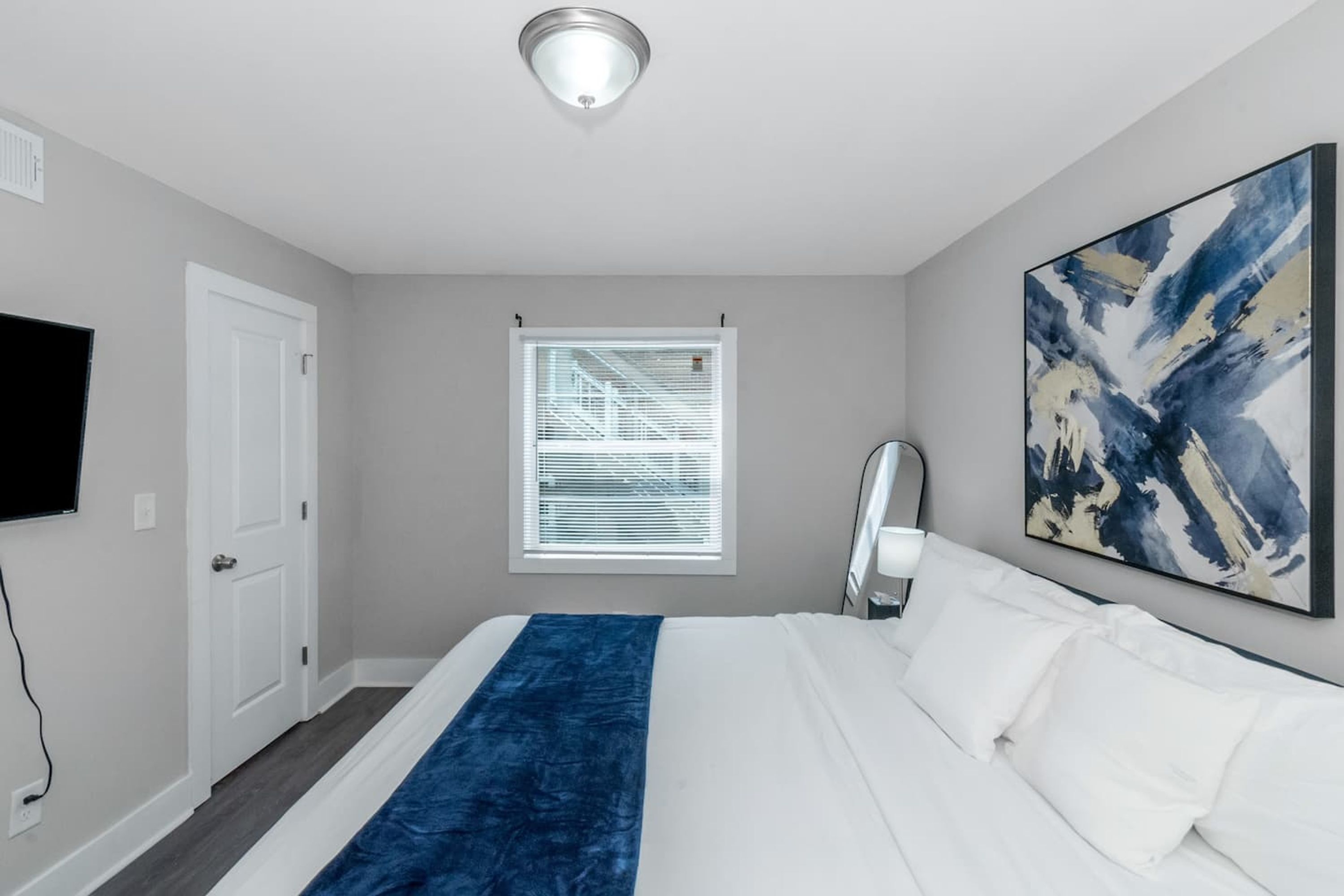 Ein Hotelzimmer mit Bett, einem großen abstrakten Gemälde, einem Fenster mit Jalousien, einer Tür und einer Deckenleuchte.