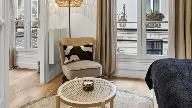 Quarto de hotel com uma cama, uma mesa redonda de madeira com tampo trançado, uma cadeira com almofada estampada, um abajur e vista da cidade através de grandes janelas com cortinas bege.