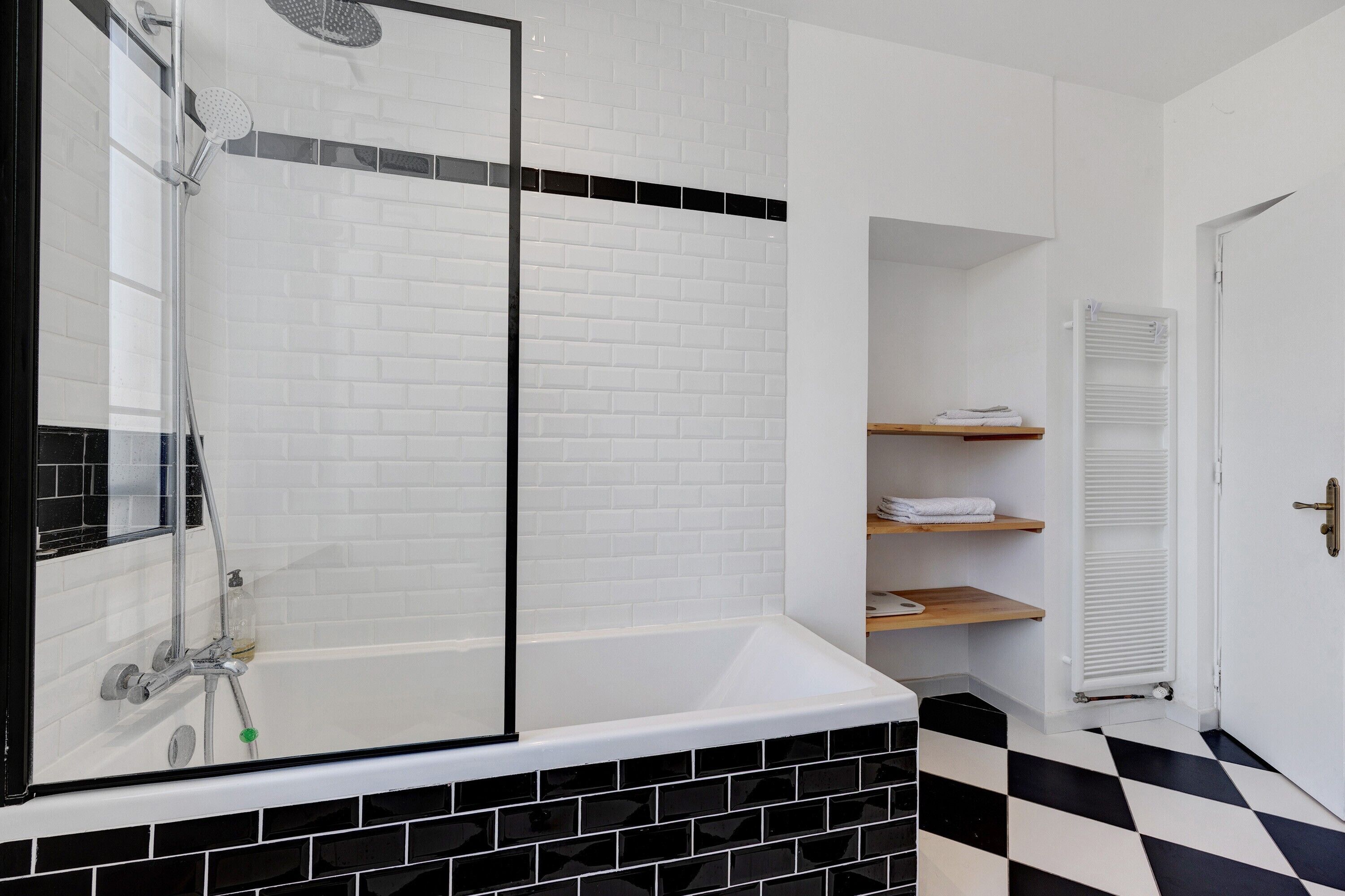 A bathroom with a glass-enclosed shower, a white tiled wall, a black and white checkered floor, and a white door.