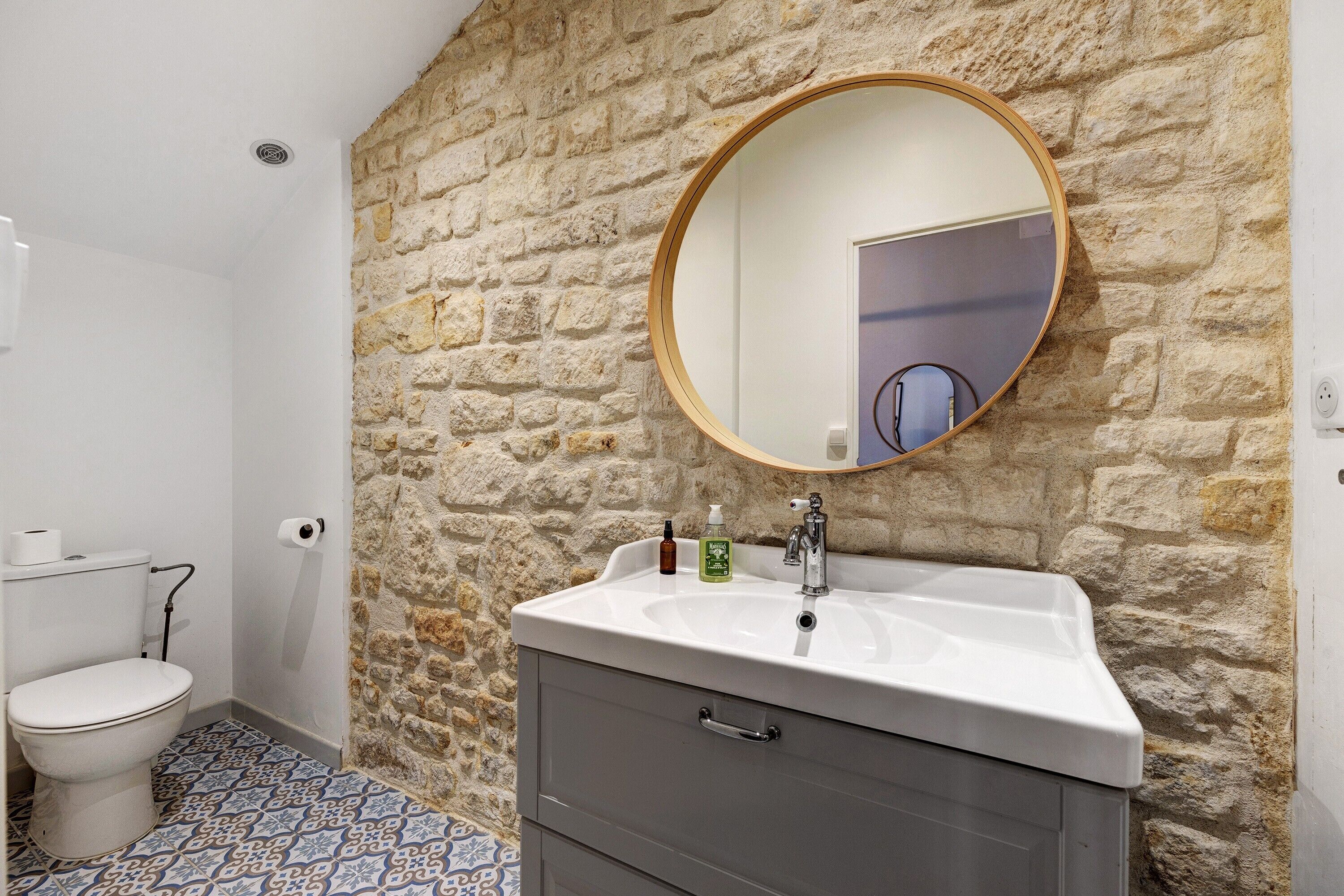 Une salle de bain avec un mur en pierre, un miroir rond, un lavabo avec un robinet et des toilettes.