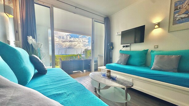 Ett modernt vardagsrum med en turkos soffa, ett soffbord i glas och ett stort fönster med utsikt över omgivningen.